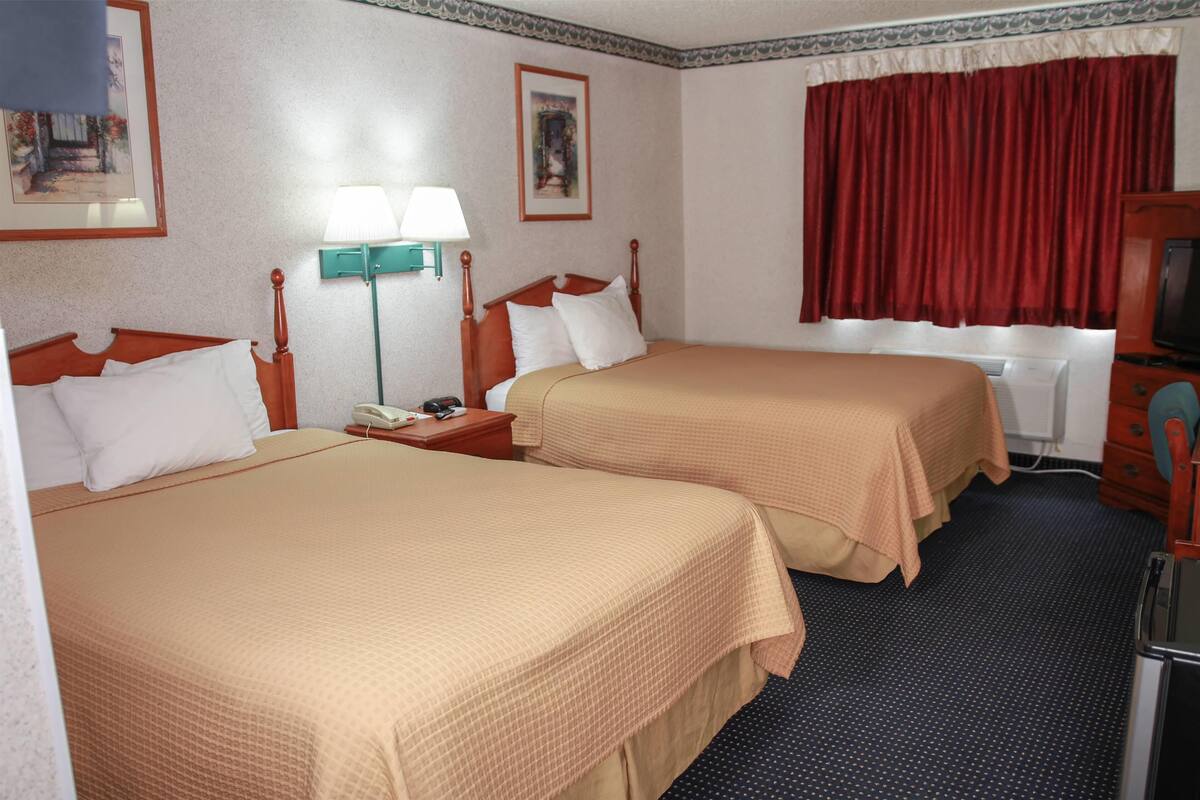 Quarto de hotel com duas camas, uma mesa com luminária, televisão e janela com cortinas vermelhas.