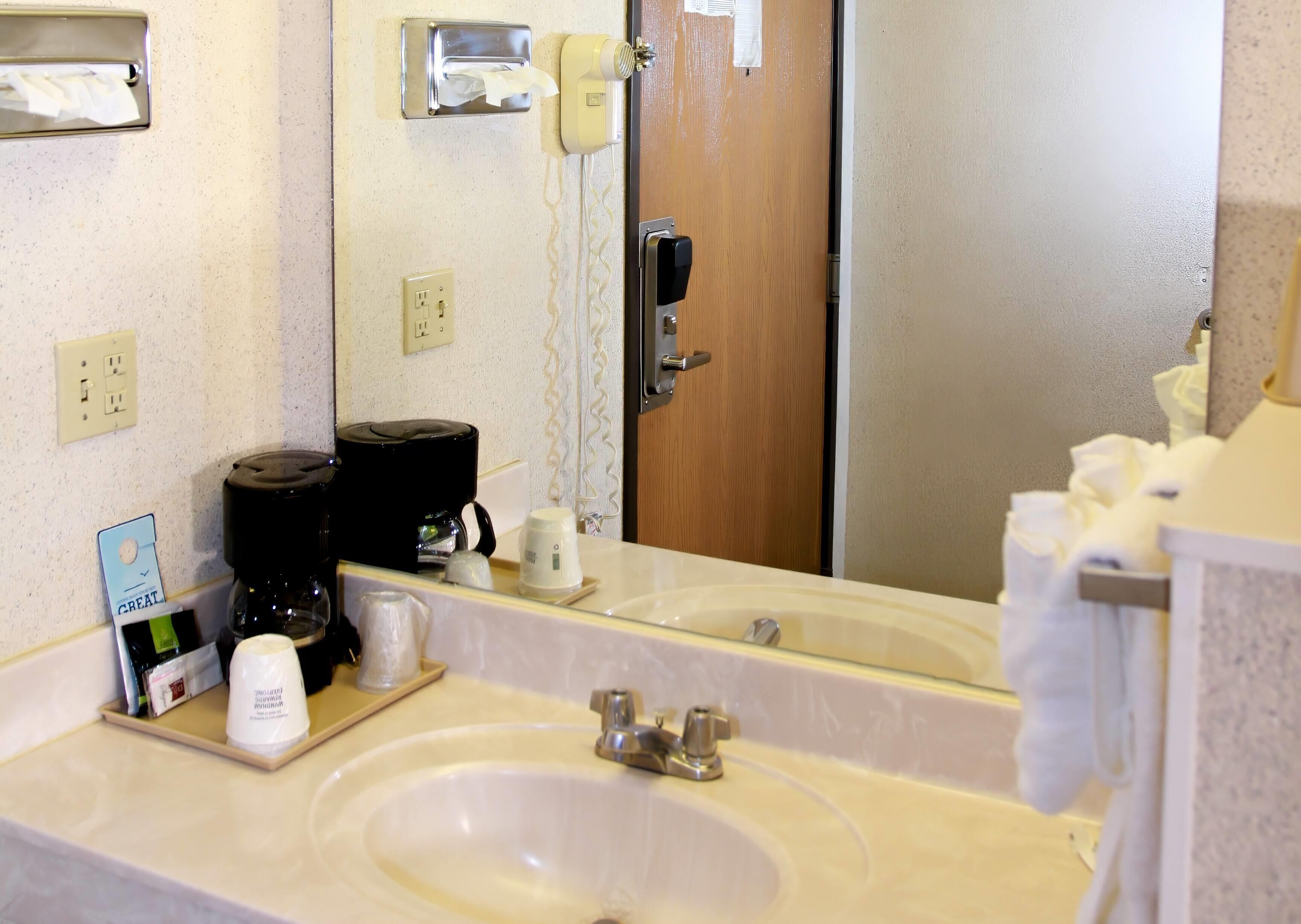 A hotel bathroom with a sink, mirror, towel dispenser, and a coffee maker.
