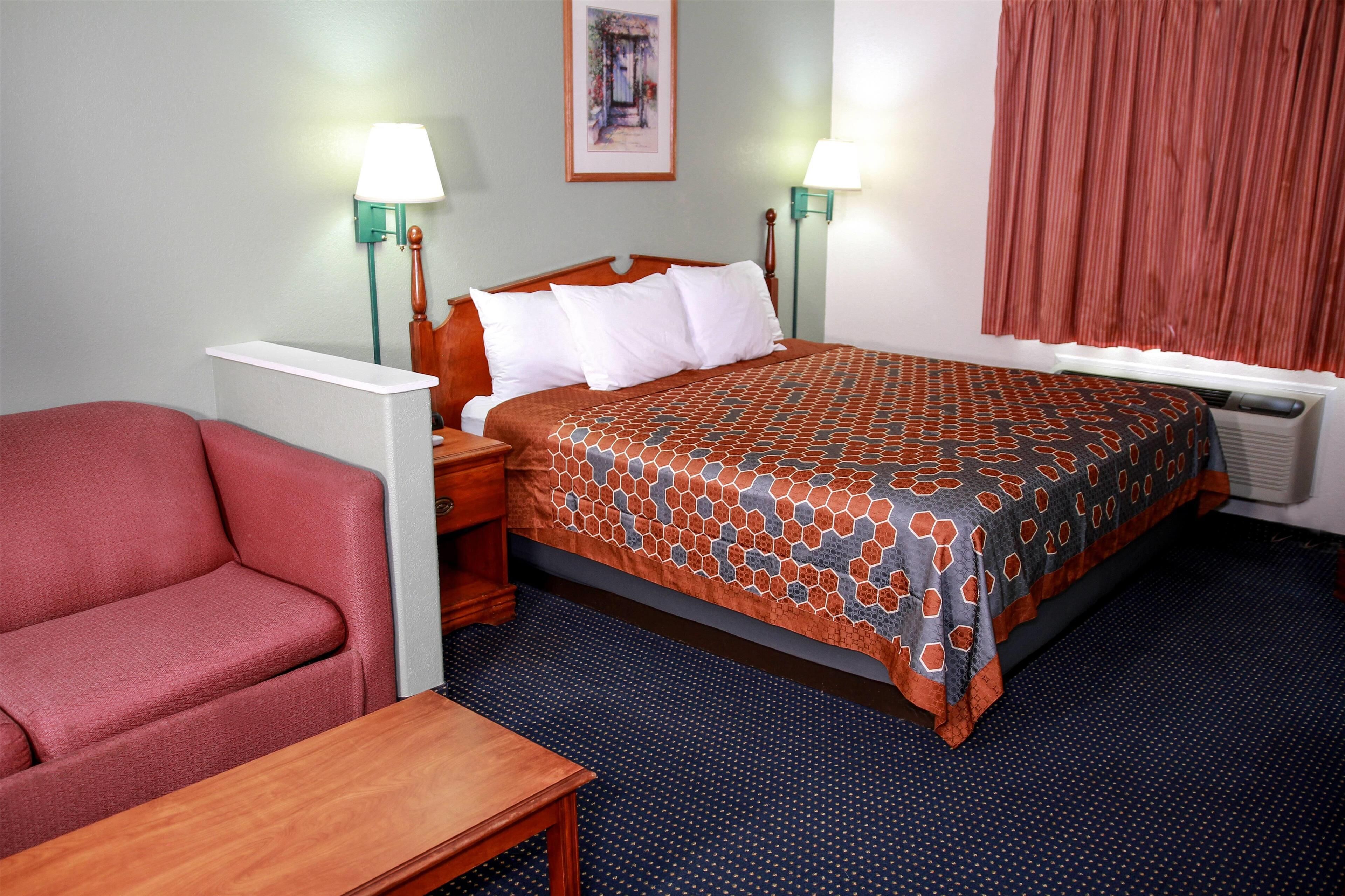 A hotel room with a bed, a red armchair, a wooden nightstand, two wall-mounted lamps, a framed picture on the wall, and a window with curtains.