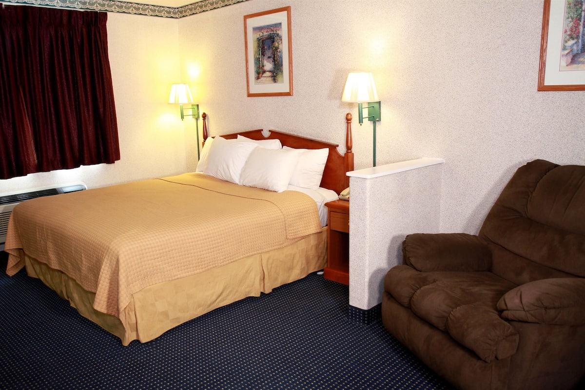 Quarto de hotel com uma cama, duas luminárias de cabeceira, uma poltrona marrom e uma mesinha.