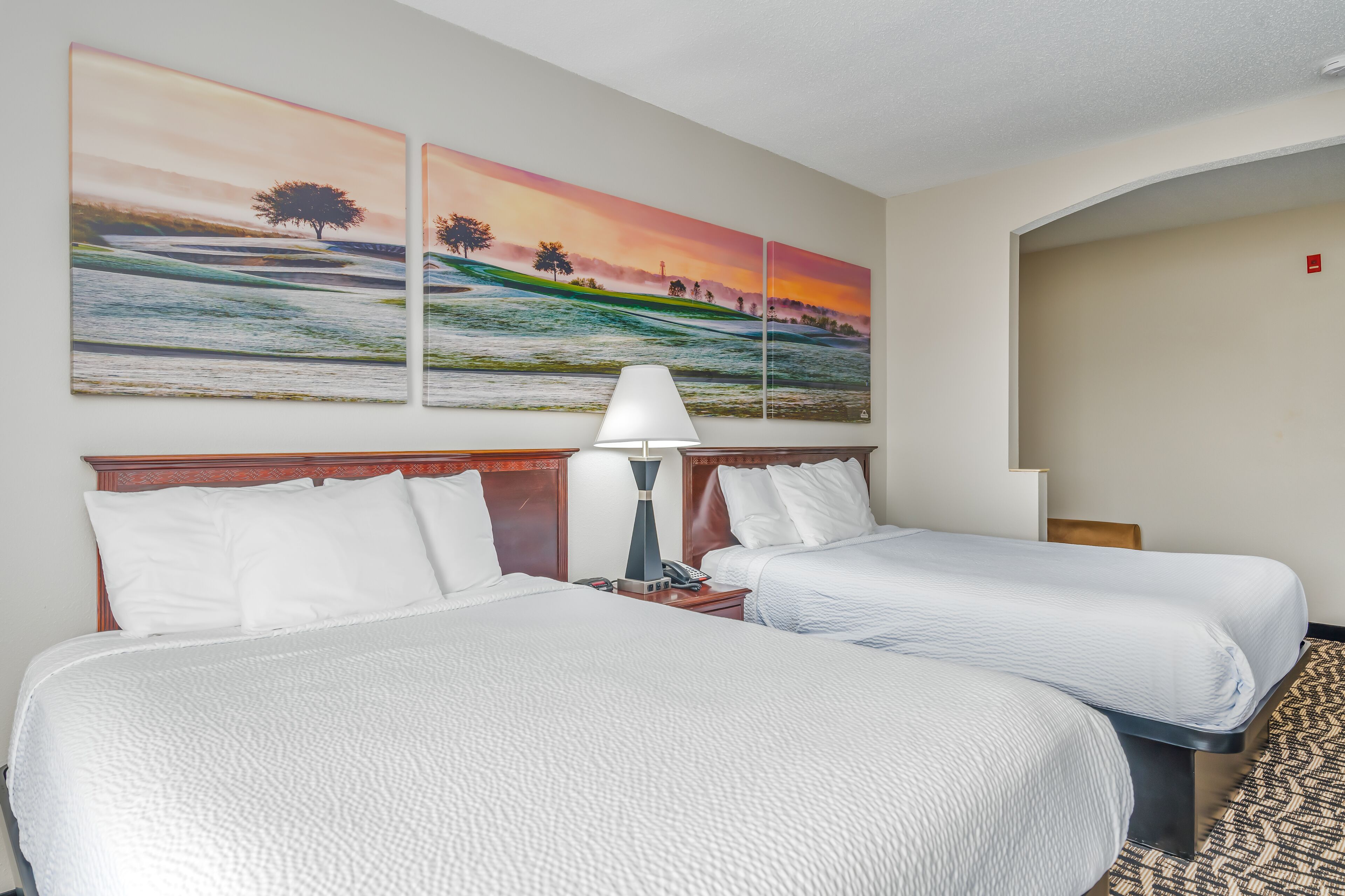 A hotel room with two beds, a nightstand, a lamp, and two paintings on the wall.