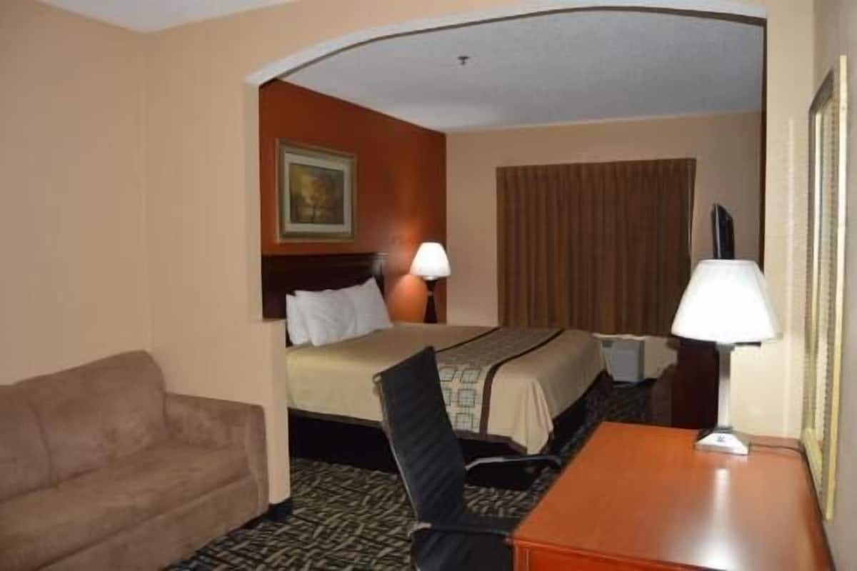 A hotel room with a bed, a desk, a chair, a lamp, a painting on the wall, and a window with curtains.