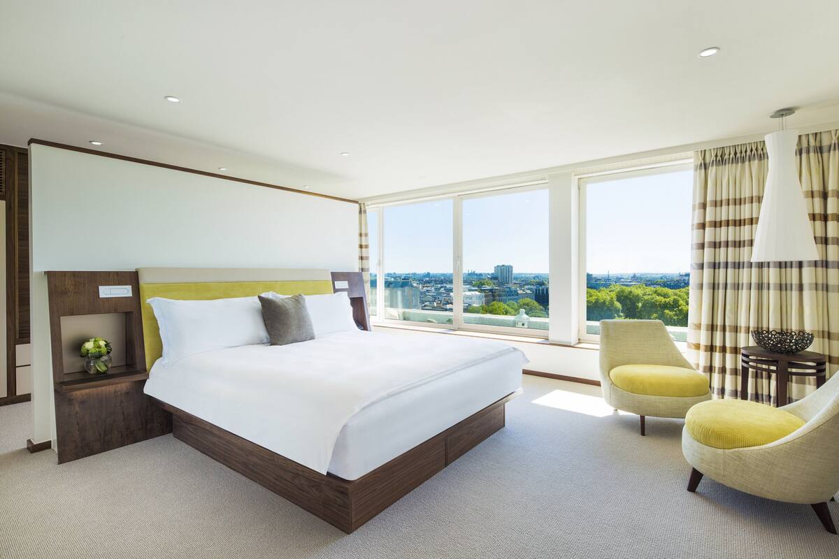 A modern hotel room with a large bed, a view of the city, and a yellow armchair.