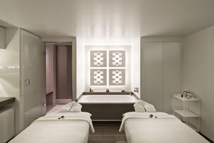 Ein modernes Hotelzimmer mit einer großen Badewanne, zwei Betten, einem Schminktisch mit Spiegel und einem Fenster mit geometrischem Muster.