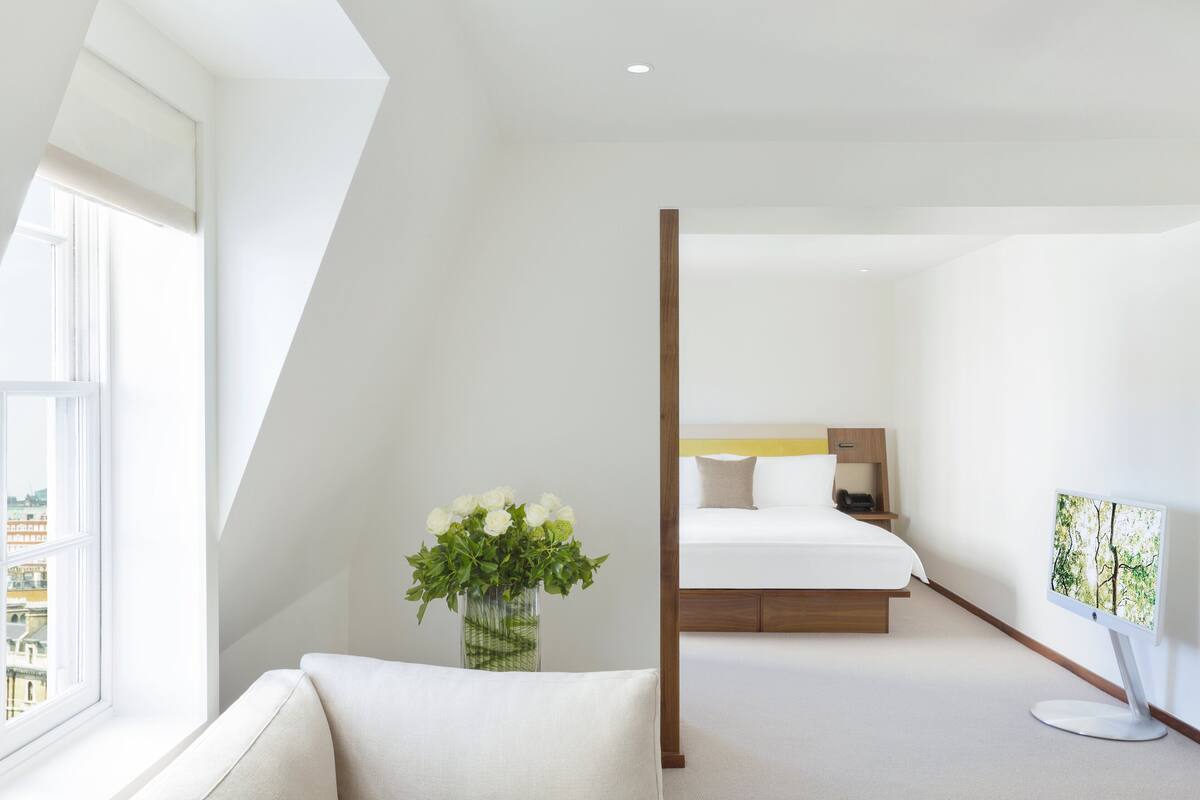 A modern hotel room with a large window, a bedside table with a vase of flowers, a bed with a wooden headboard, and a flat-screen TV on the wall.