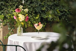 A table with a white tablecloth, a clear glass vase with flowers, and a cocktail glass with fruit on a white plate.