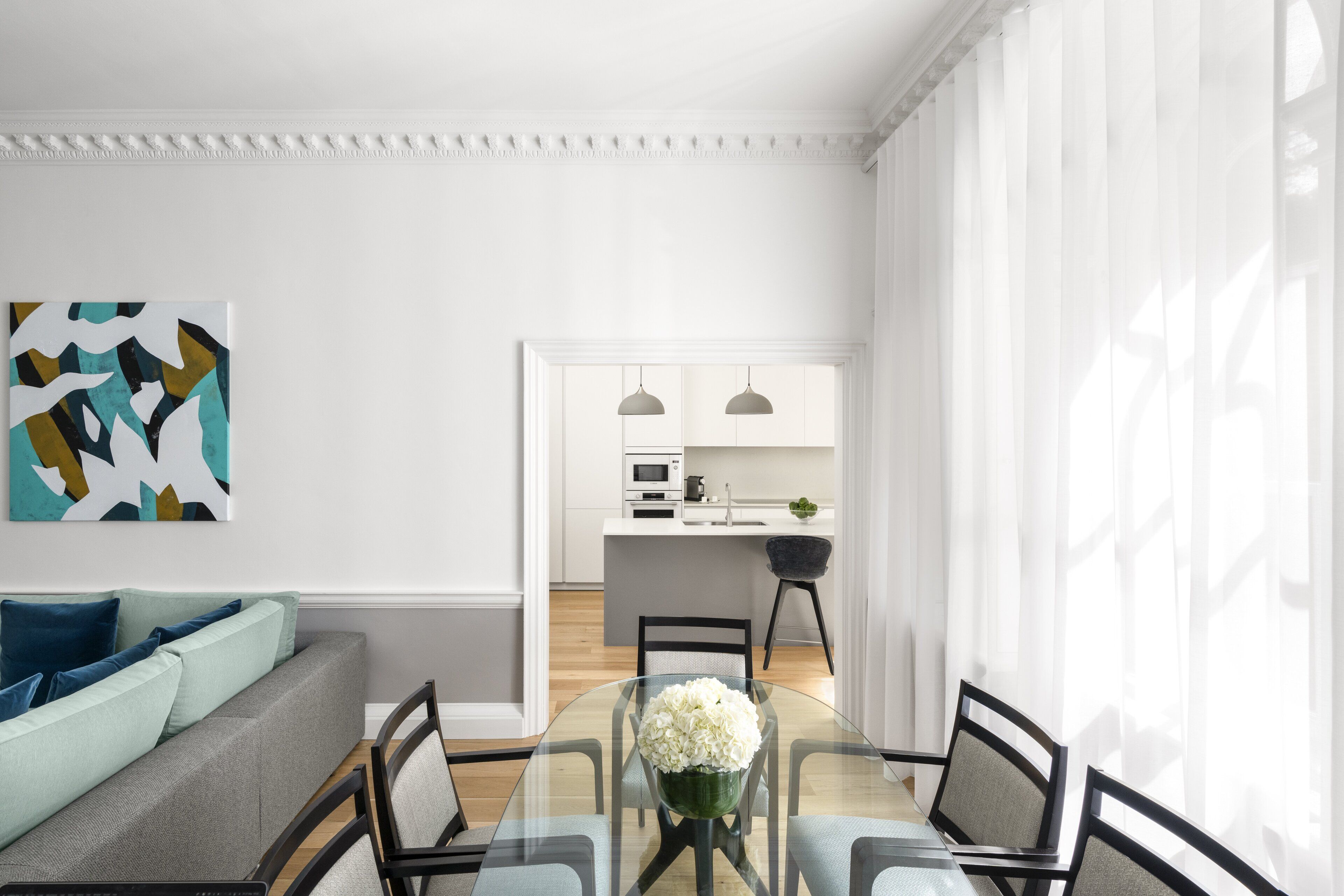 A modern living room with a glass dining table, a sofa with cushions, and a kitchen in the background.