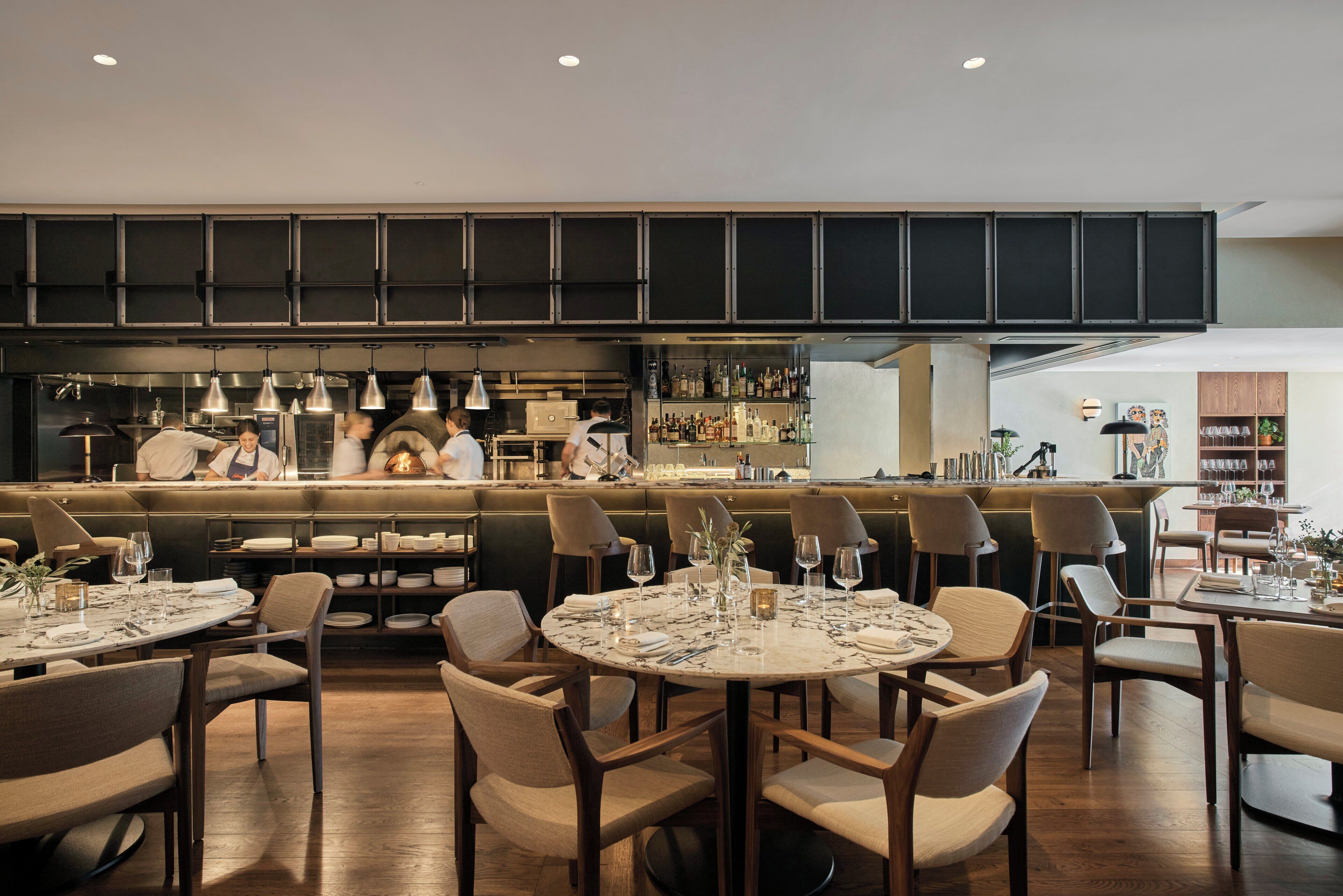 A modern restaurant with round tables, chairs, and a bar area with a kitchen in the background.