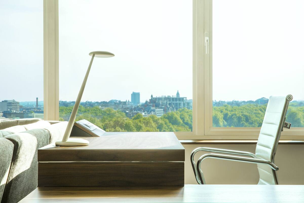 A modern office chair and desk with a laptop, a tablet, and a smartphone. A large window offers a view of a cityscape and greenery.