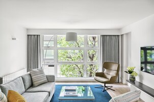 A modern living room with a sofa, armchair, and coffee table, large windows with curtains, and a view of trees.