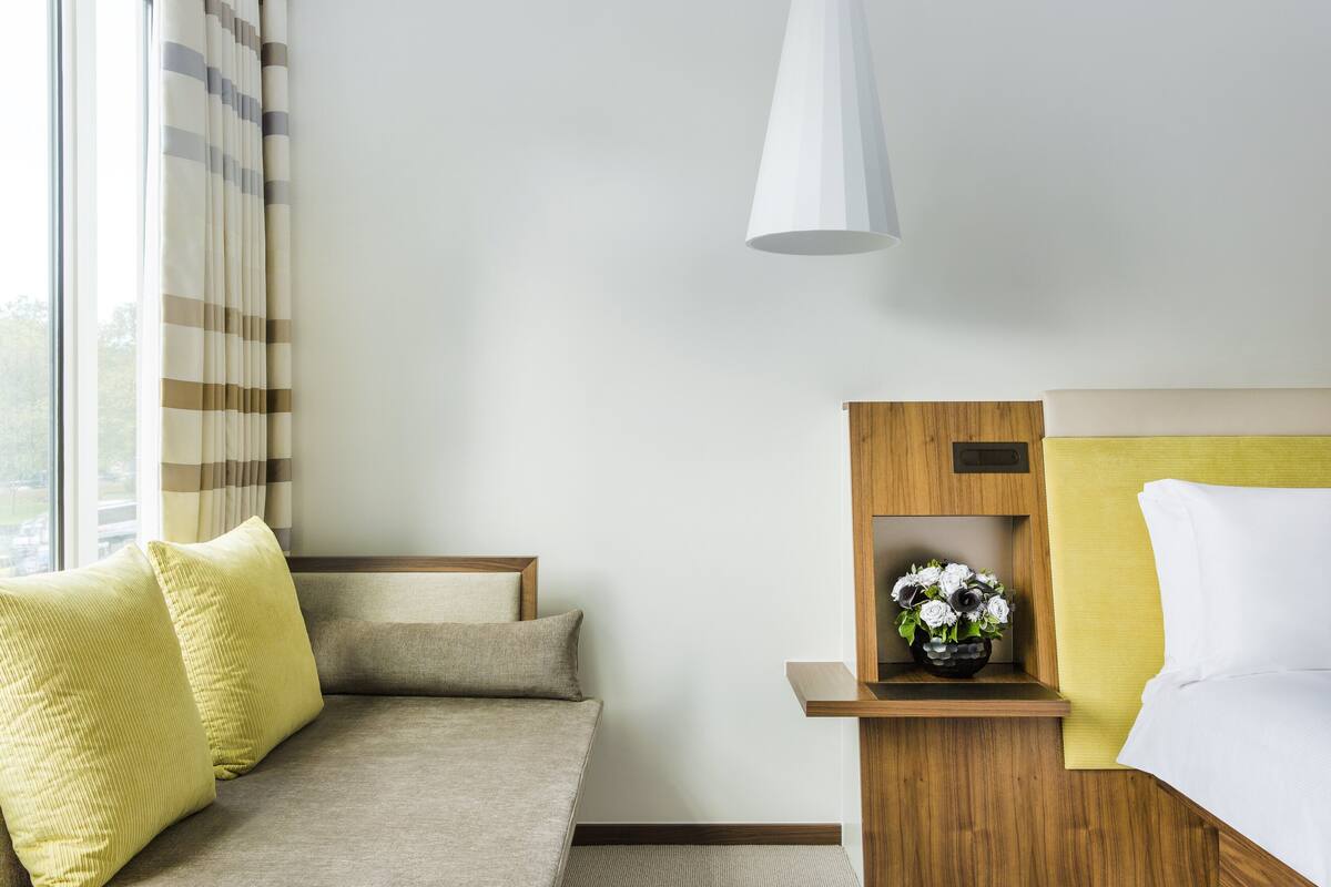 A hotel room with a sofa, a bed, a bedside table with a vase of flowers, and a hanging light fixture.