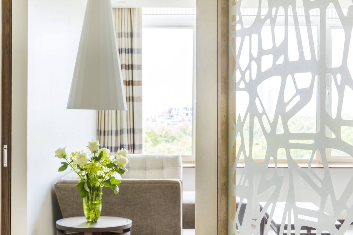 A room with a white chair, a round table with a vase of flowers, a lamp, and a view through a window with curtains.