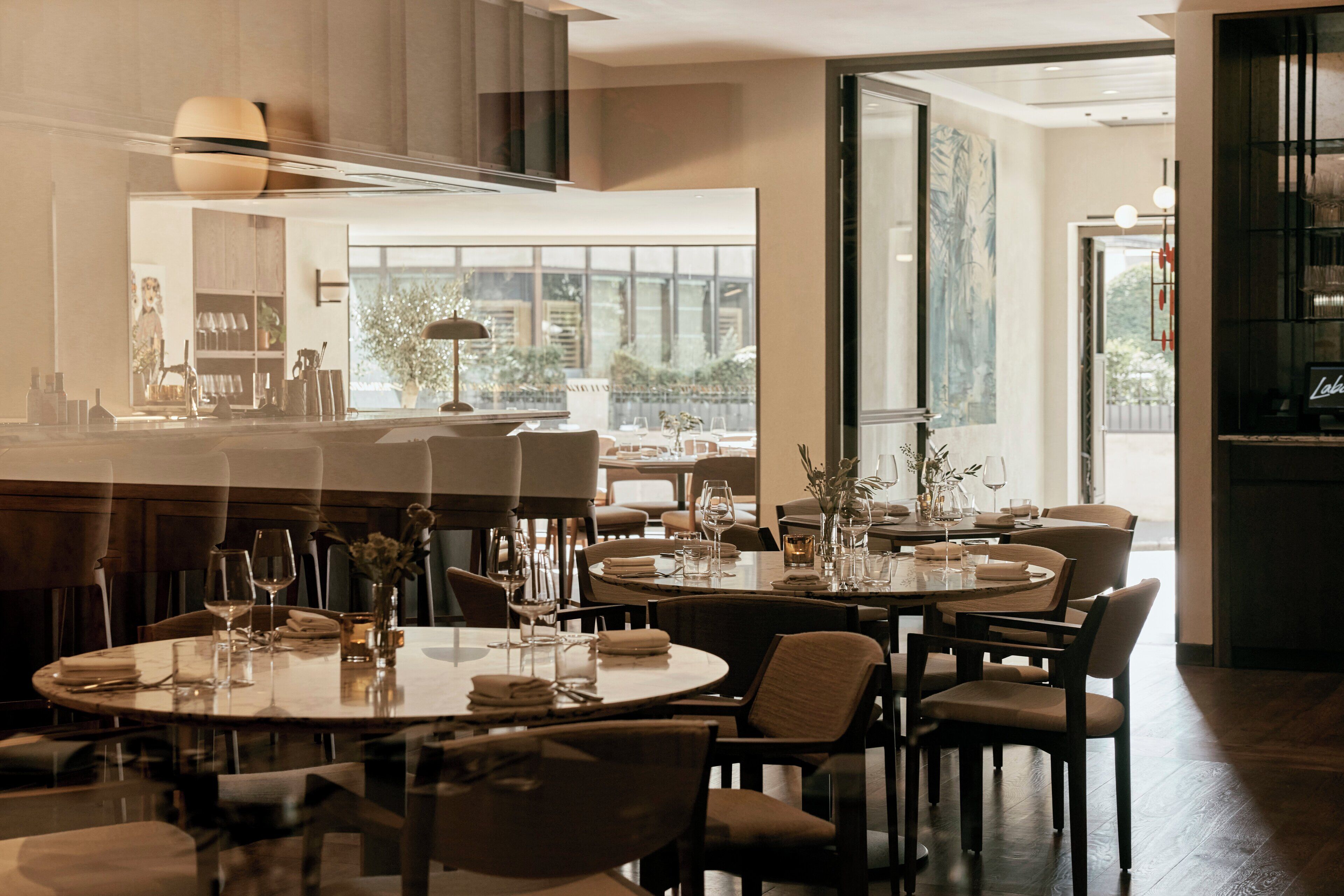 A modern dining area with round tables and chairs, a large window offering a view of the outdoors, and a kitchen visible in the background.