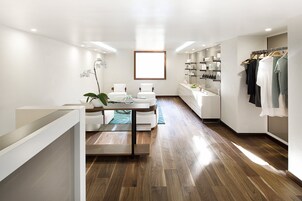 Ein modernes, helles Zimmer mit Holzboden, einem Glastisch mit Stühlen, einem weißen Sofa, einem großen Fenster und einem Kleiderständer mit verschiedenen Kleidungsstücken.