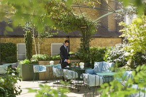 Eine Außenpatio-Terrasse mit Tischen und Stühlen, ein Kellner, der Speisen serviert, und ein Backsteinhaus im Hintergrund.