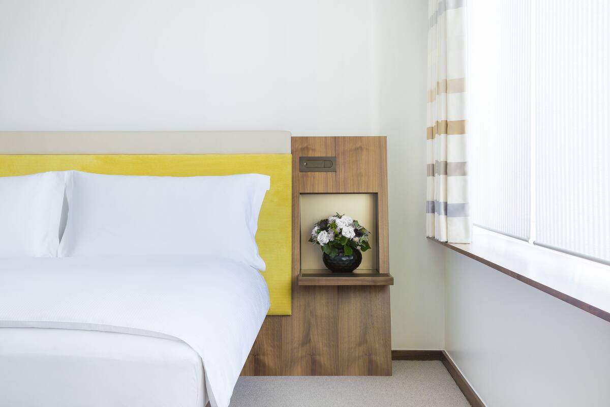 A hotel room with a bed, a small wooden nightstand with a vase of flowers, and a window with blinds.