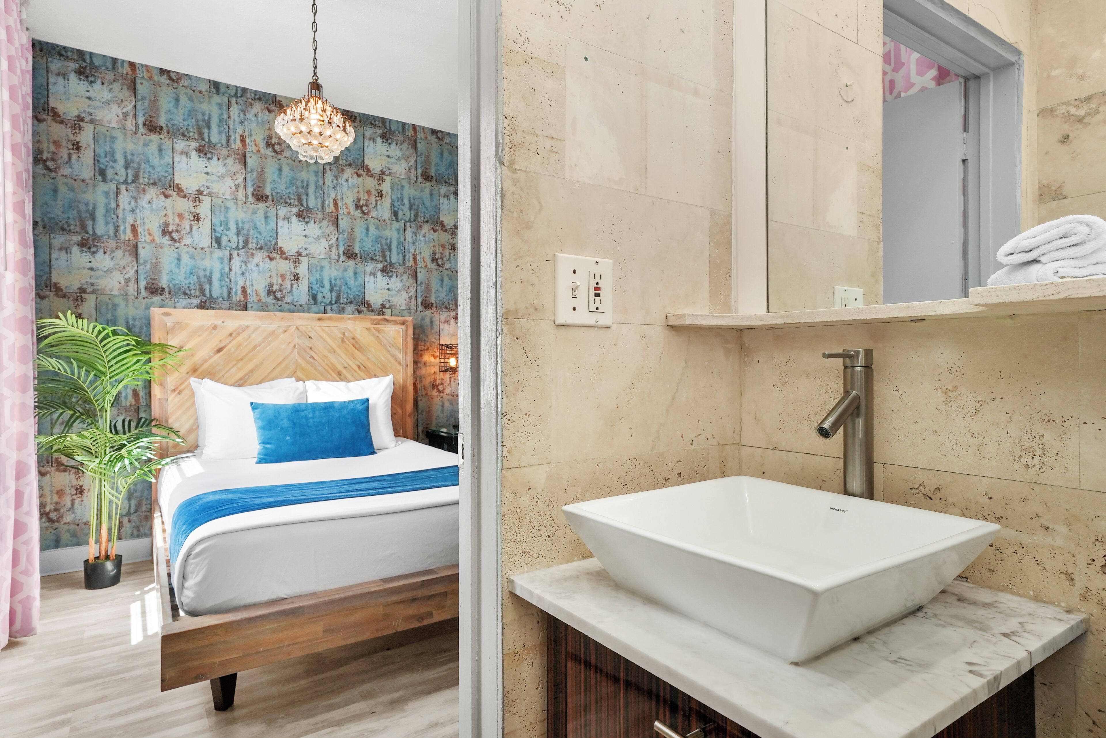 A modern bedroom with a wooden headboard, a large bed, a bedside plant, and a bathroom with a white sink and marble countertop.