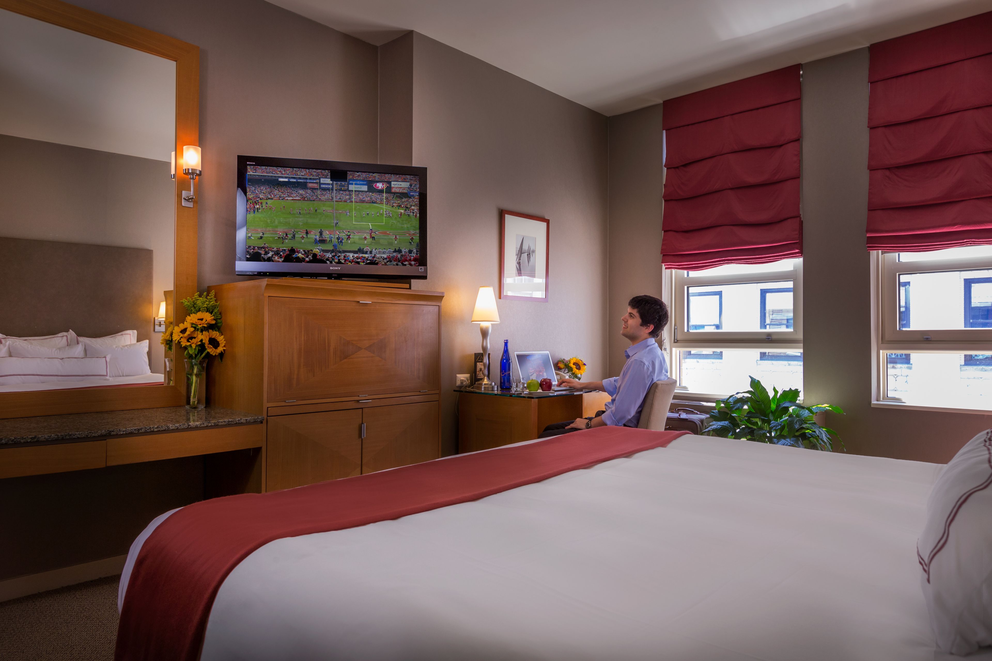 Een hotelkamer met een groot bed, een aan de muur bevestigde televisie, een bureau met een lamp en een raam met rode gordijnen.