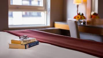 A hotel room with a bed, a window with a view, a lamp, and a desk with a flower arrangement.