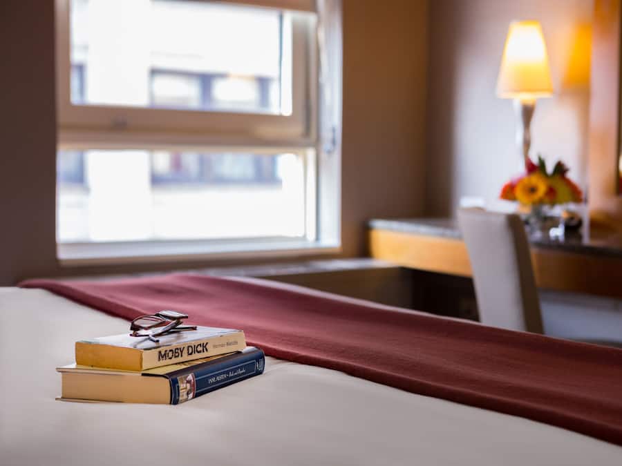 A hotel room with a bed, a window with a view, a lamp, and a desk with a flower arrangement.
