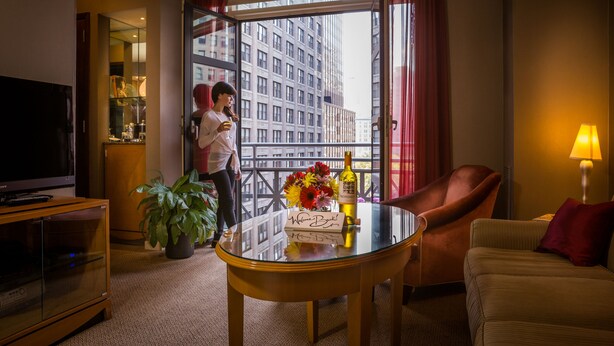 Habitación de hotel con una mesa de vidrio, sofá, televisor, planta en maceta y balcón con vistas a la ciudad.