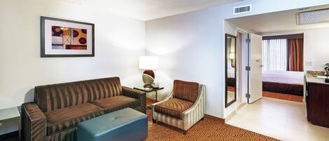 A hotel room with a brown sofa, a blue ottoman, a striped armchair, a small table with a lamp, and a framed picture on the wall.