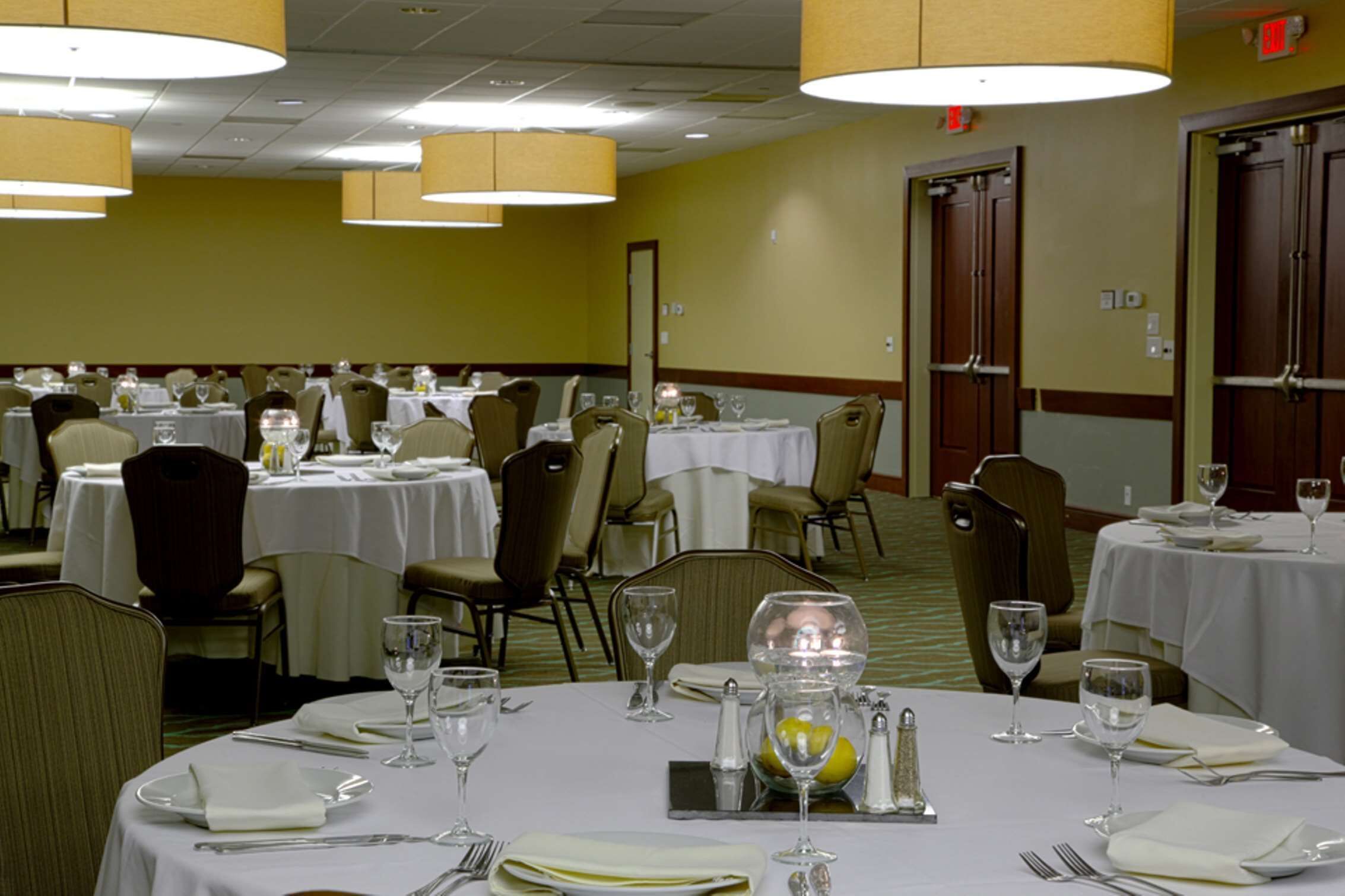 Un salón de conferencias con varias mesas redondas, cada una con mantel blanco, sillas y lo necesario para comer, como platos, vasos y cubiertos.