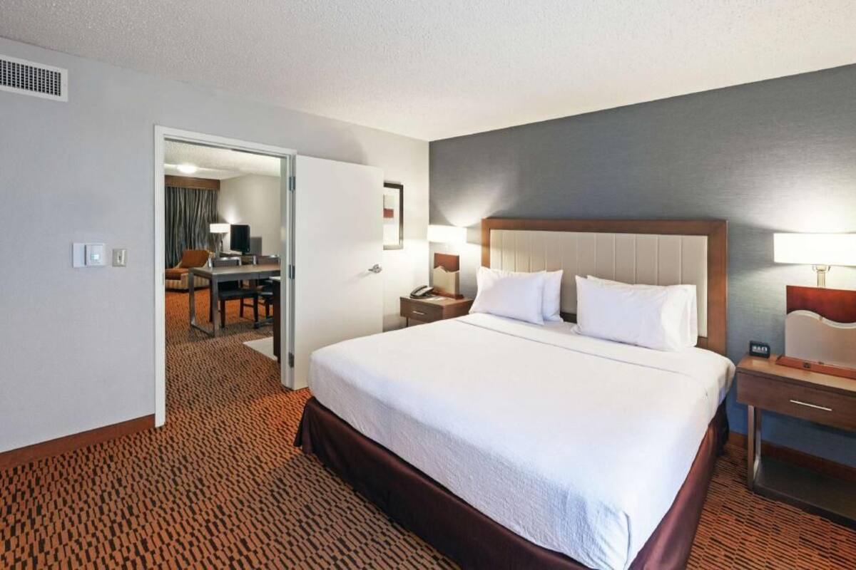 A hotel room with a large bed, bedside tables, a desk with a chair, and a door leading to another room.