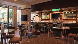 A bar area with multiple seating options, including round tables and bar stools. There is a television mounted on a stone pillar, and the bar is stocked with various bottles and glasses. - Embassy Suites by Hilton Dallas Market Center (Dallas)