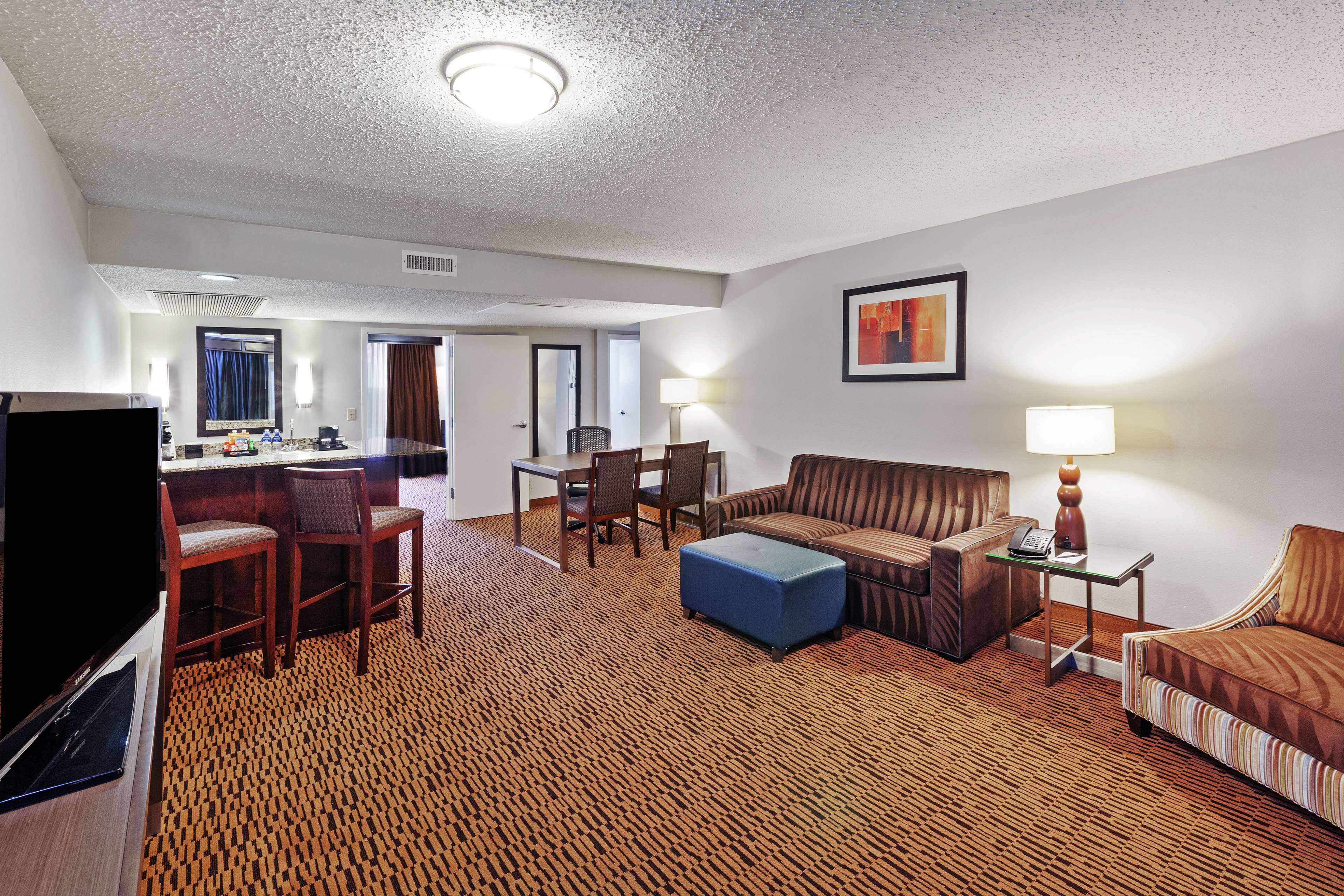 A hotel room with a living area, a dining table with chairs, a sofa, a TV, and a lamp.