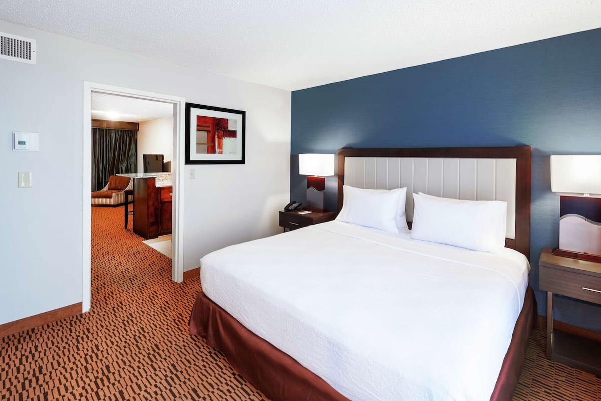 A hotel room with a large bed, two bedside tables with lamps, a door leading to a sitting area, and a framed picture on the wall.