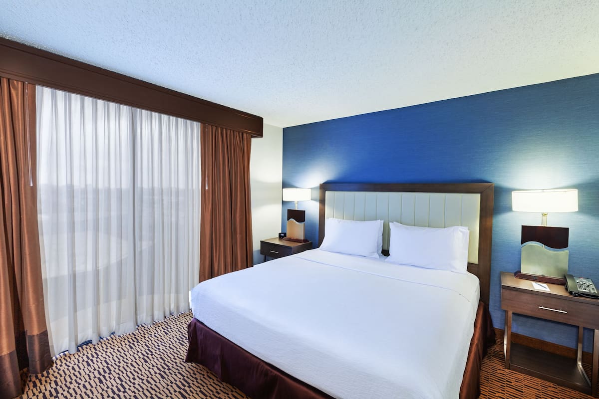 A hotel room with a large bed, a nightstand with a lamp, a telephone, and a window with curtains.