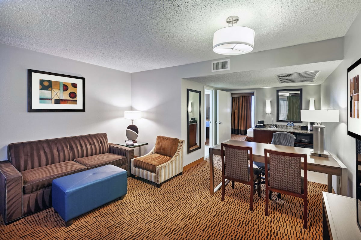 A hotel room with a sofa, armchair, ottoman, dining table with chairs, kitchenette, and a large mirror.