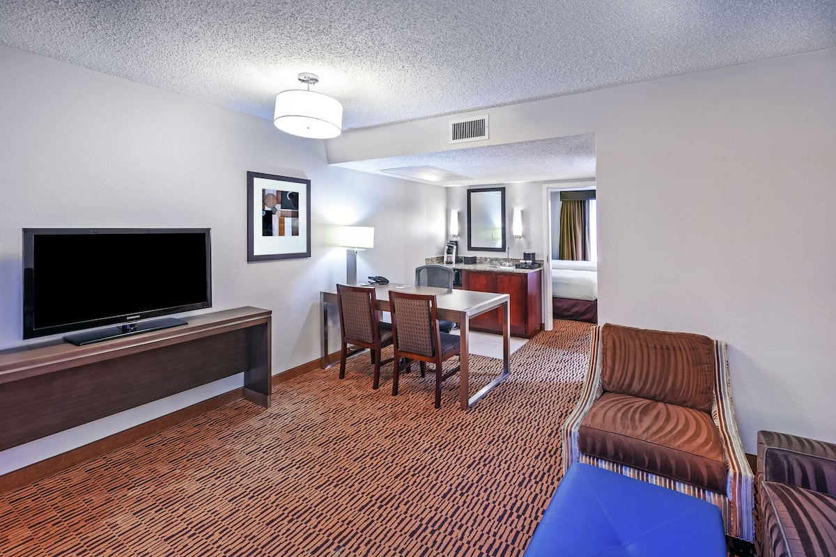 A hotel room with a flat-screen TV, a dining area with chairs and a table, a sofa, and a small kitchenette.