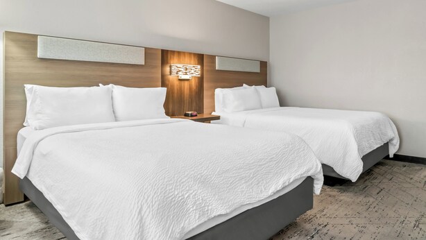 A hotel room with two beds, a wooden headboard, a wall-mounted light fixture, and a patterned carpet. - Holiday Inn Express Lapeer by IHG (Lapeer)