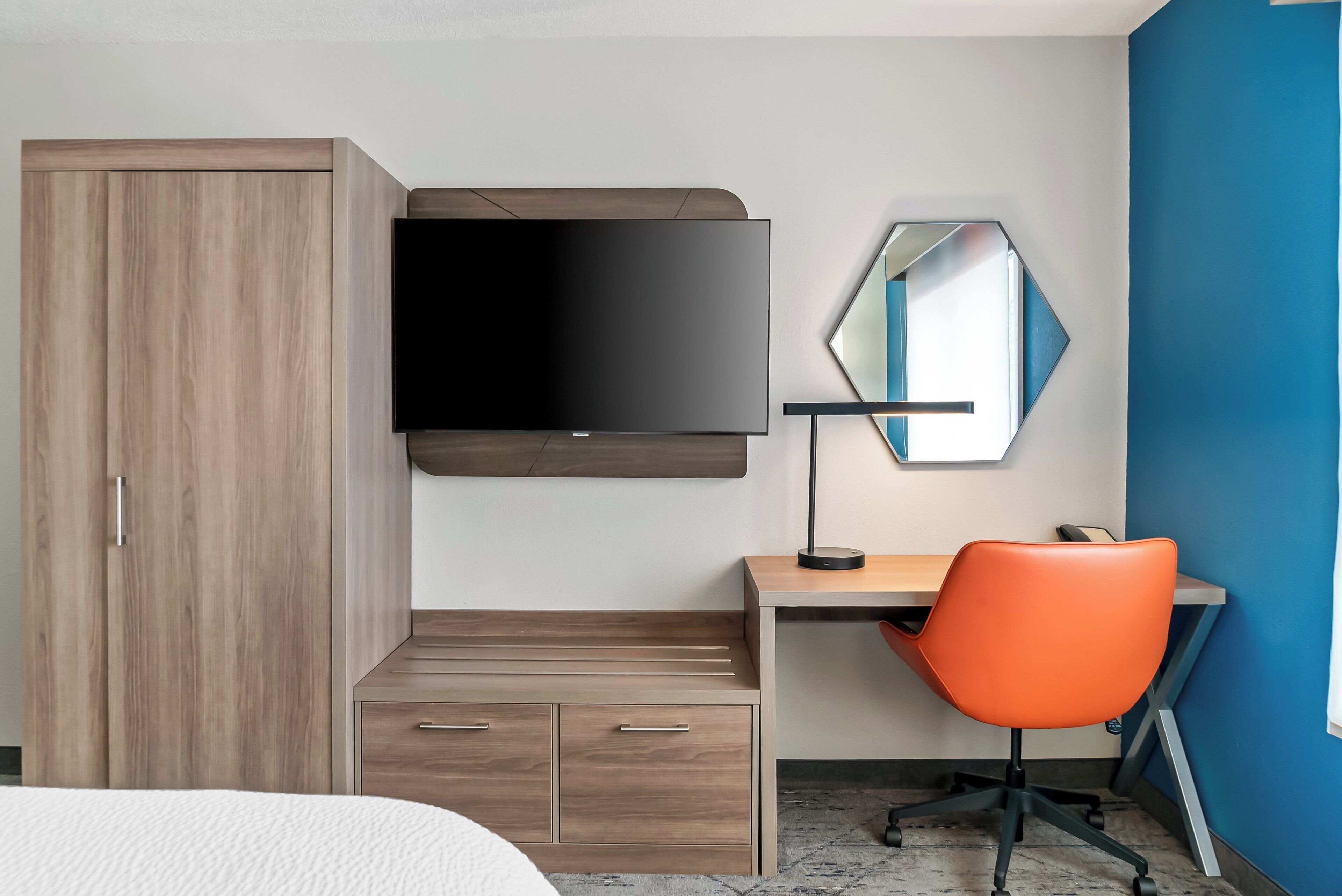 A modern hotel room with a wooden wardrobe, a flat-screen TV mounted on the wall, a desk with a chair, and a mirror.