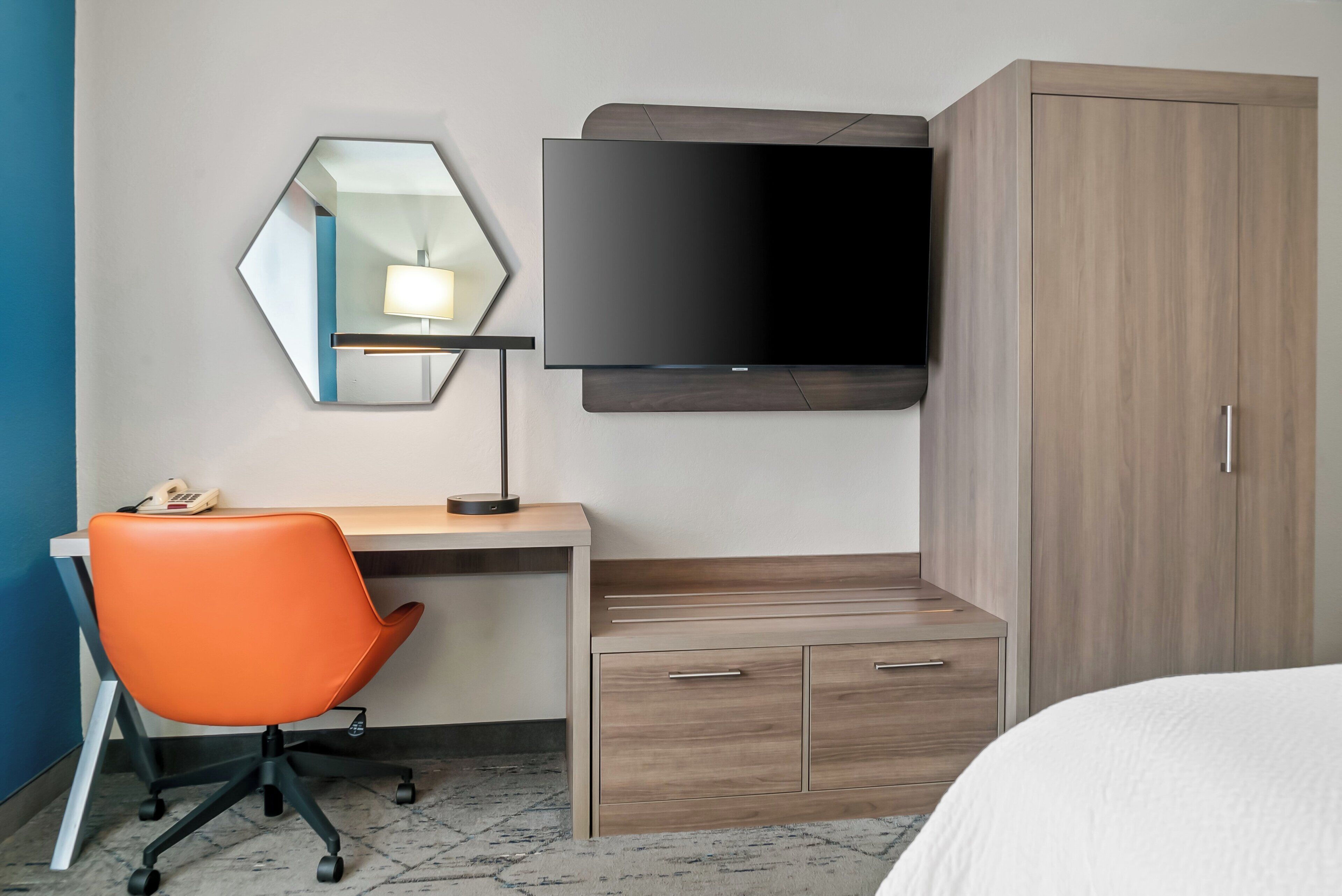 Una habitación de hotel con escritorio, una silla de oficina naranja, un televisor de pantalla plana montado en la pared, un armario de madera y una cama con ropa blanca.
