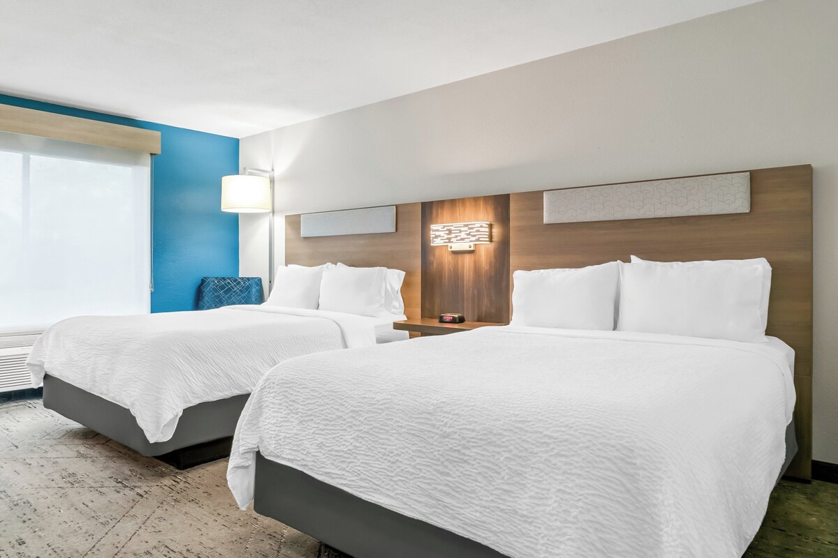 A hotel room with two beds, a large window, a wall-mounted lamp, and a wooden headboard.