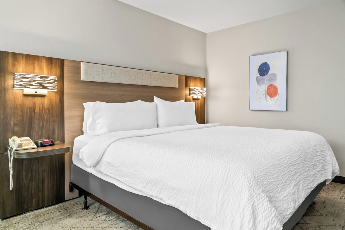 A modern hotel room with a large bed, wooden headboard, bedside lamps, a telephone, and a framed abstract painting on the wall.