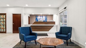 A reception area with two blue armchairs, a wooden coffee table, and a reception desk with a blue and white design. - Holiday Inn Express Lapeer by IHG (Lapeer)