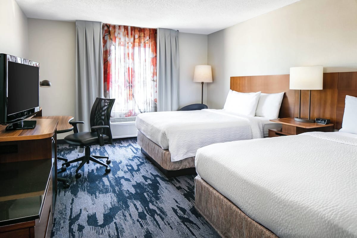 A hotel room with two beds, a desk with a TV, a chair, a lamp, and a window with curtains.