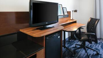 Habitación de hotel con escritorio de madera, una silla de oficina negra, televisor de pantalla plana, un refrigerador pequeño negro y una lámpara.