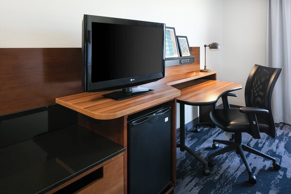 Une chambre d’hôtel avec un bureau en bois, un fauteuil de bureau noir, un téléviseur à écran plat, un petit réfrigérateur noir et une lampe.