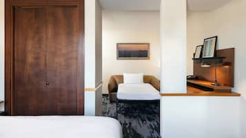 Habitación de hotel con una puerta grande de madera, una cama con edredón blanco, un sofá con almohada blanca, un escritorio pequeño con lámpara y un cuadro en la pared.