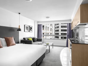 A modern hotel room with a bed, sofa, dining table, kitchenette, and large windows with a city view. - Mantra on Kent (Sydney)