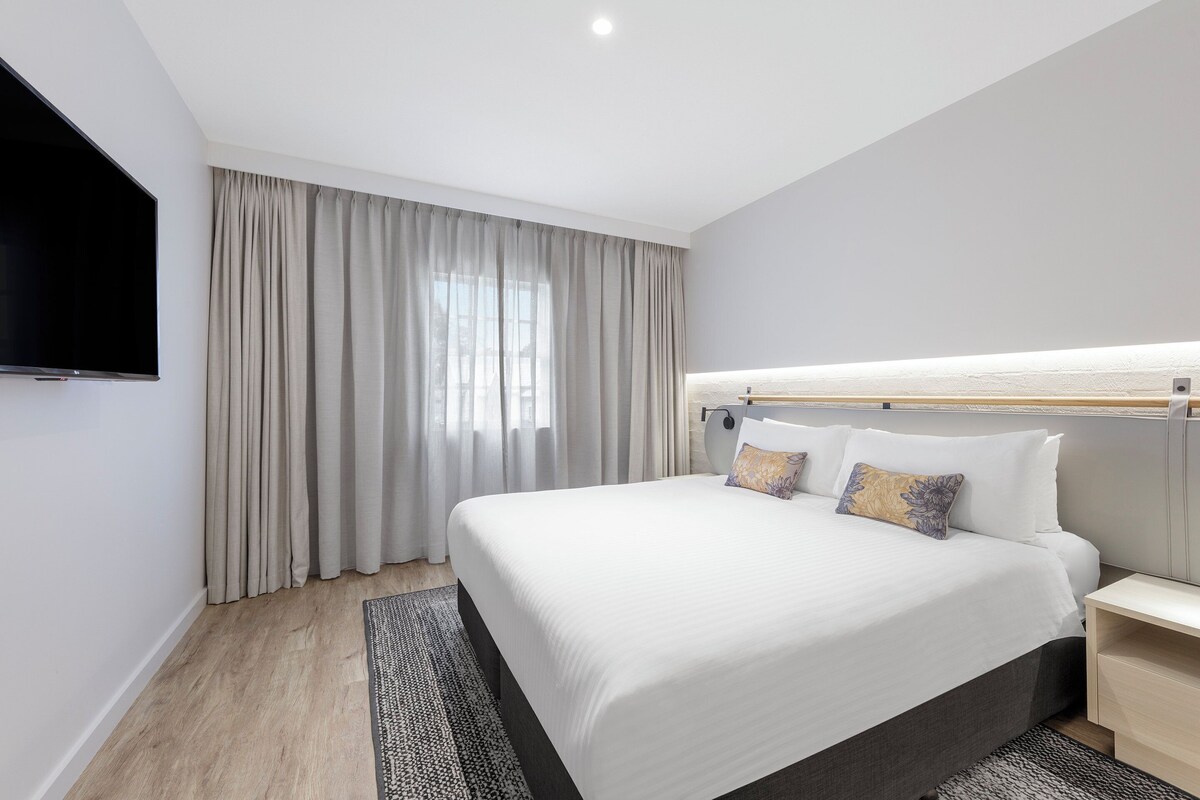 A modern hotel room with a large bed, a flat-screen TV, a bedside table, and a window with curtains.
