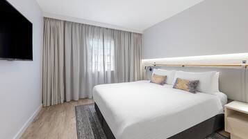 A modern hotel room with a large bed, a flat-screen TV, a bedside table, and a window with curtains.