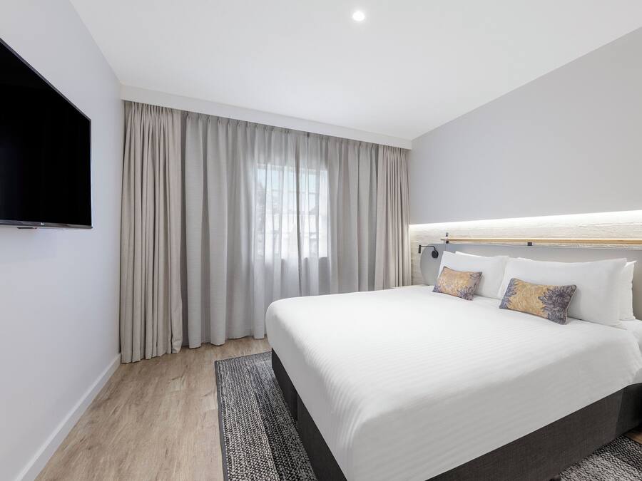 A modern hotel room with a large bed, a flat-screen TV, a bedside table, and a window with curtains.