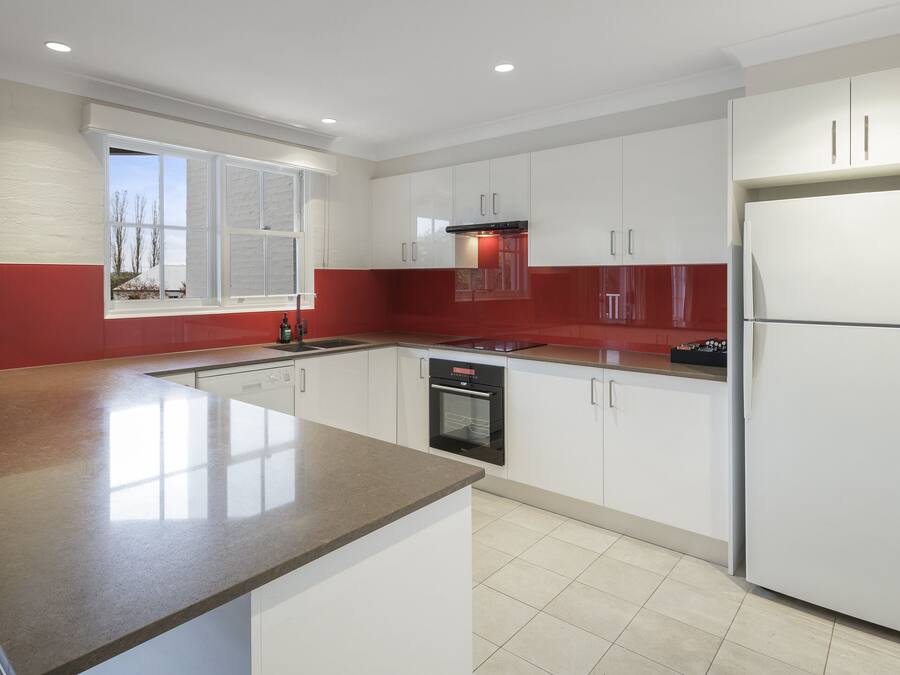 A modern kitchen with white cabinets, a red accent wall, a large island, and a built-in oven.