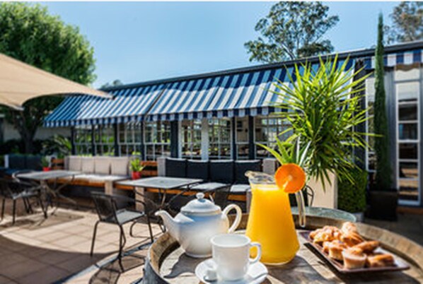Un café en plein air avec des tables, des chaises, une théière, un pichet de jus d’orange et un plateau de pâtisseries.
